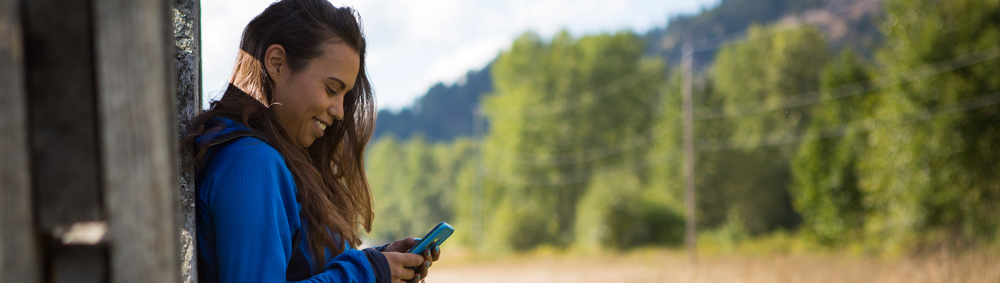 A young woman in a blue jacket is leaning against a building near a field and looking at her phone and smiling.